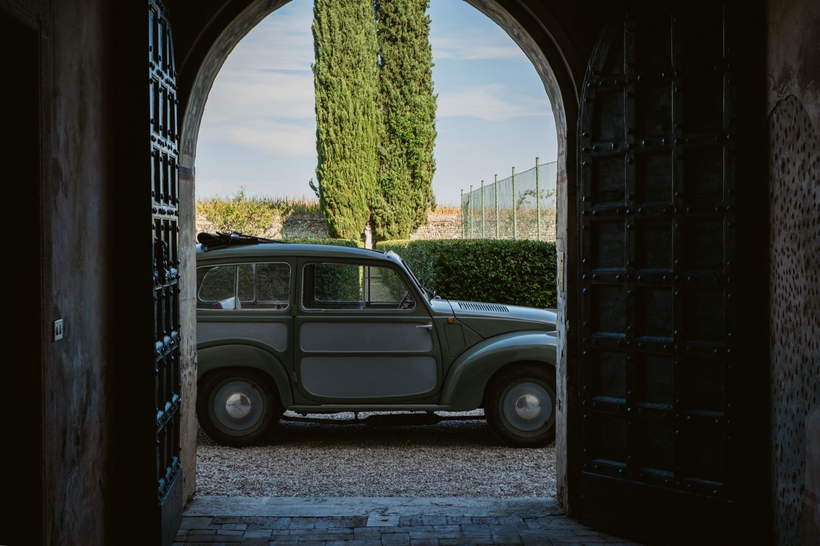A vintage car beyond the open gate, among the cypresses