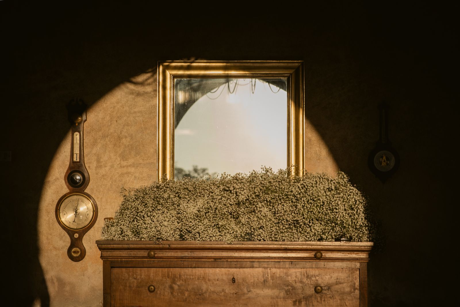 A gilded mirror above a chest of drawers covered in baby’s breath, in the afternoon light